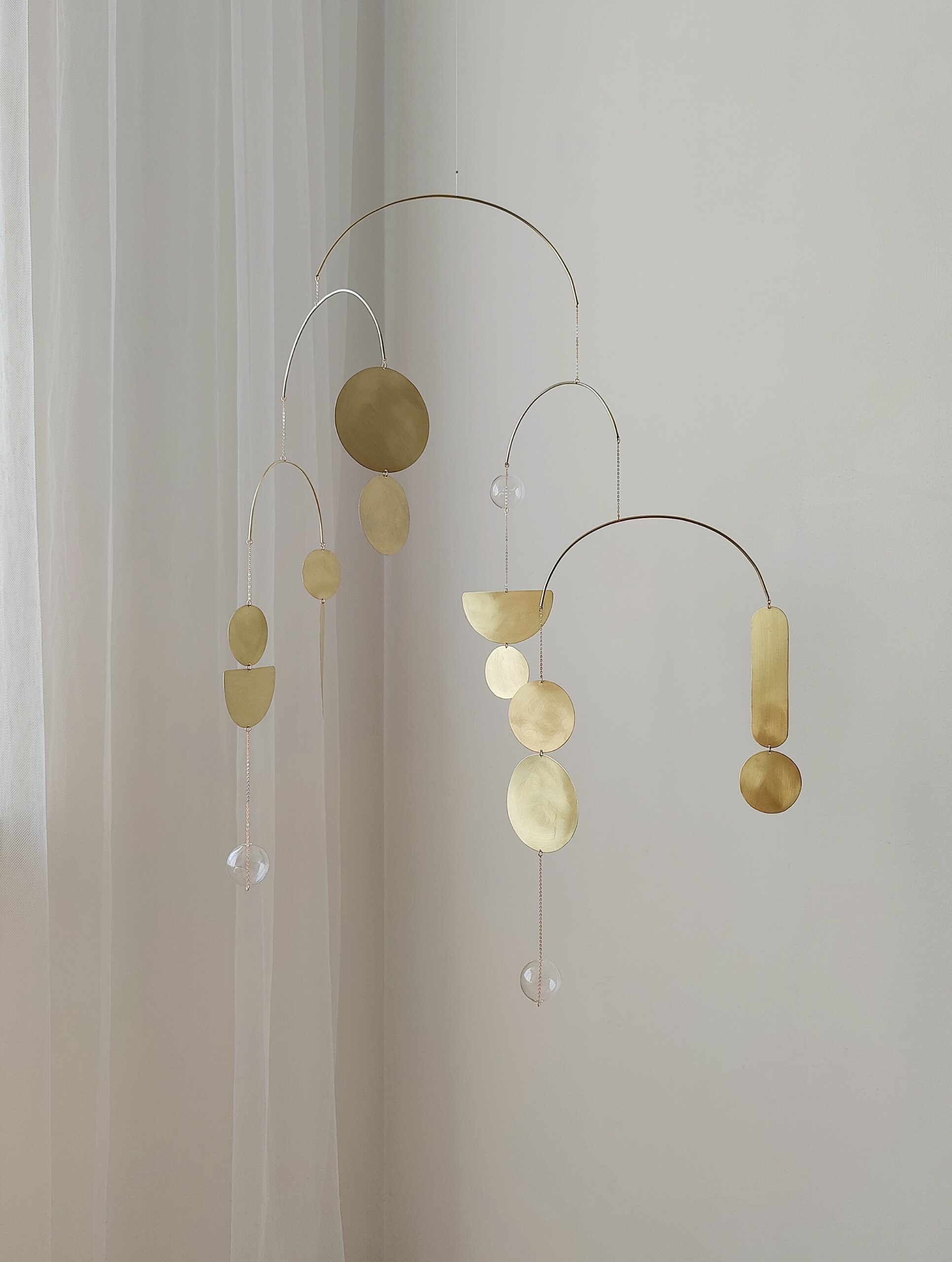 Elegant gold kinetic mobile with circular, semicircular, crescent moon shapes and glass balls hangs in front of white wall. Mobile has 5 thin brass interconnected arches. This hanging brass sculpture casting soft shadows on the light wall. Serene ambiance.