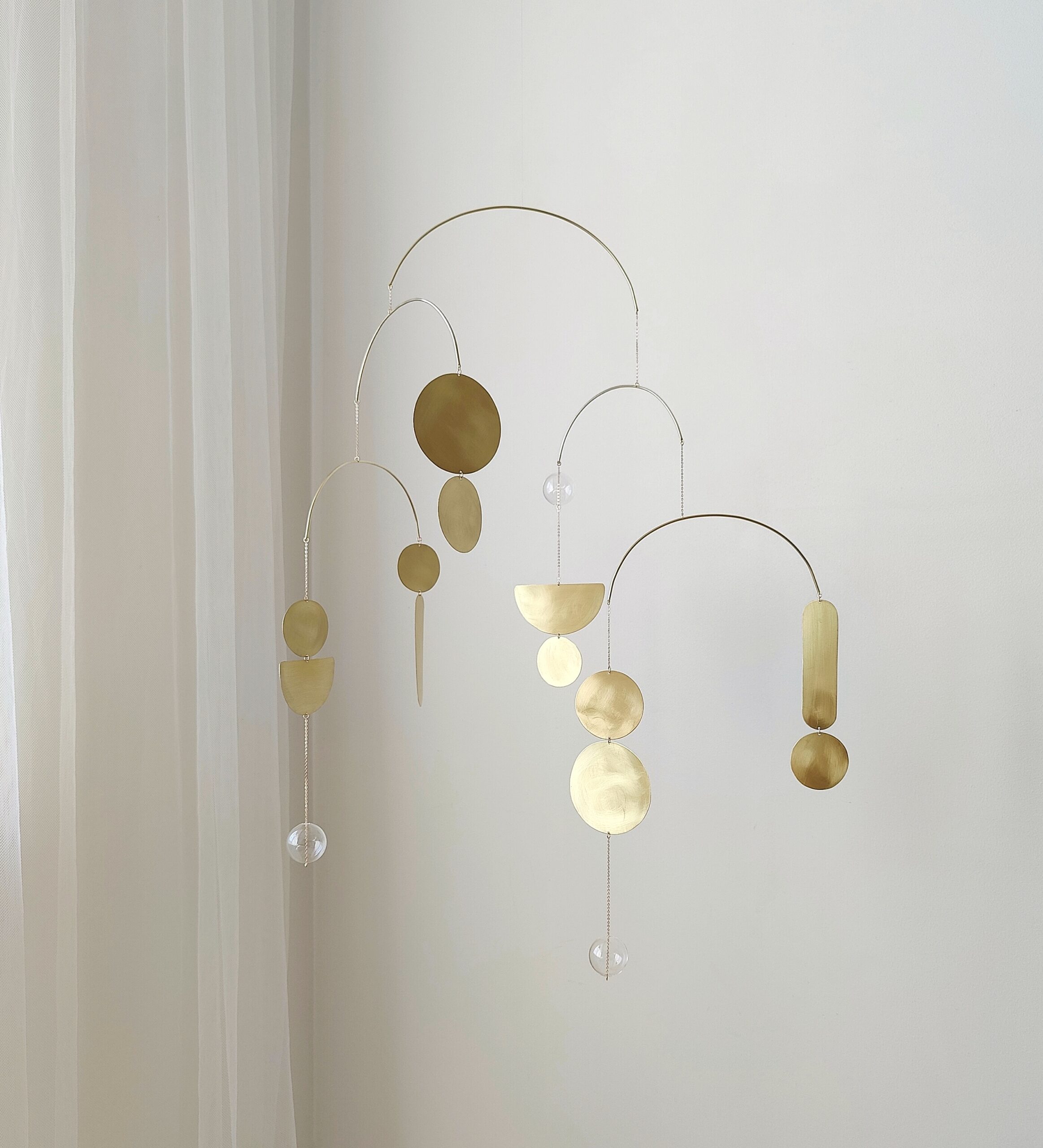 Elegant gold kinetic mobile with circular, semicircular, crescent moon shapes and glass balls hangs in front of white wall. Hanging brass sculpture casting soft shadows on the light wall. Serene ambiance.