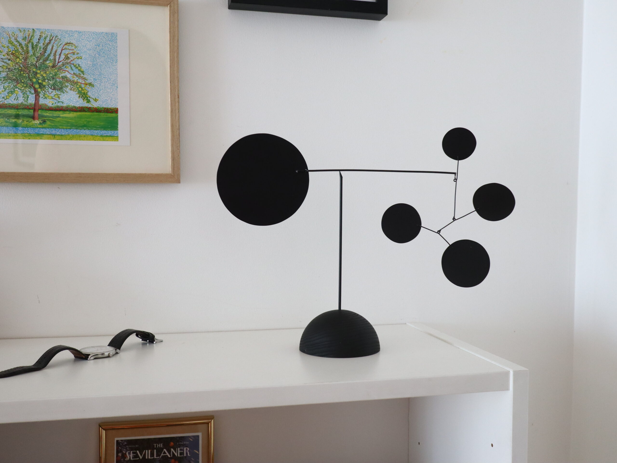 A metal black abstract mobile sculpture with circular elements is displayed on a white shelf